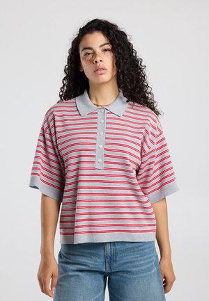 Jonge vrouw met krullend donker haar, gekleed in een grijs poloshirt met korte mouwen met rode strepen en een blauwe spijkerbroek, staand tegen een effen achtergrond.