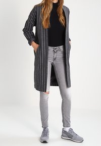 Cárdigan largo a rayas grises con abertura en la parte delantera, combinado con una camiseta negra y jeans rotos en gris claro. Unos zapatos deportivos grises completan el conjunto.