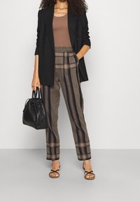 Svart dubbelknäppt blazer över en brun ribbad topp, tillsammans med bruna mönstrade byxor. Tillbehör inkluderar en svart väska och sandaler.