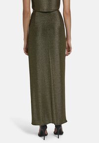 Langes, figurbetontes Kleid aus dunkelgrünem Metallicstoff, mit einem Schitz hinten, schlanker Form und passenden schwarzen High Heels.