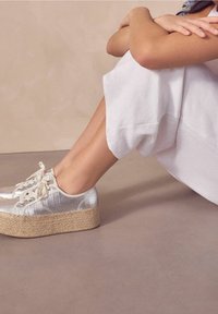 Baskets à plateforme métalliques argentées avec semelles en jute tressé, dotées d'une fermeture à lacets et d'une tige texturée. Portées avec un pantalon blanc ample.
