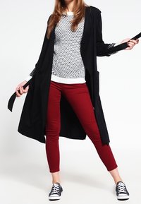Cardigan nero lungo sopra un maglione bianco a motivo e pantaloni rossi aderenti, abbinati a scarpe da ginnastica nere e bianche. Texture lisce e colori a contrasto.