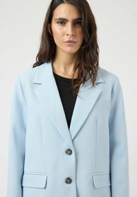 Γυναίκα με μακριά καστανά μαλλιά, φοράει ανοιχτό γαλάζιο σακάκι πάνω από μαύρο τοπ, κοιτάζει μπροστά με ουδέτερη έκφραση σε απλό φόντο.