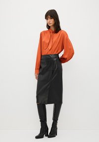 Oranžová hedvábná blůza s detailem na zavazování, kombinovaná s černou koženou midi sukni. Modelka má na sobě černé kozačky na vysokém podpatku do kolen. Jednoduchý, elegantní design.