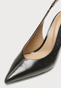 Chaussure à talon haut slingback en cuir noir à bout pointu, doublure intérieure beige et coutures visibles au niveau des orteils.