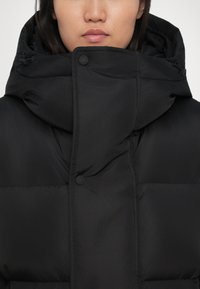 Abrigo acolchado negro con un cuello alto, que cuenta con una cremallera frontal y grandes secciones infladas. Textura suave y ligeramente brillante con un hardware mínimo.