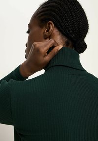 Eine Person mit geflochtenem Haar richtet den Kragen eines dunkelgrünen, gerippten Rollkragenpullovers aus, von der Seite und von hinten gezeigt.