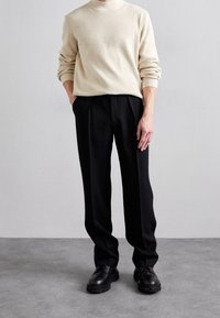 Beige Rippstrick-Turtleneck-Pullover, schwarze maßgeschneiderte Hose und klobige schwarze Schuhe. Einfaches Design, lockere Passform und glatte Texturen.