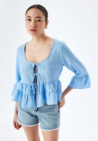 Lichtblauw peplumtop met een vierkant decolleté, strikdetail aan de voorkant en driekwartbelmouwen. Gemaakt van een текстuurstof. Draag met denim shorts.