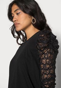 Top preto com mangas em renda de malha texturizada e detalhes em babados. O corpo é liso, criando um contraste de materiais e designs.