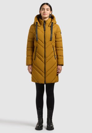 Donna che indossa un cappotto imbottito con cappuccio color senape con cerniera nera, leggings neri e stivaletti neri, in piedi davanti a uno sfondo neutro.