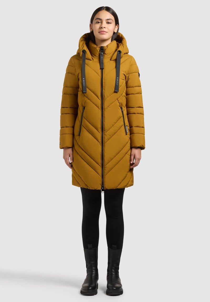 Femme portant un manteau matelassé à capuche jaune moutarde avec une fermeture éclair noire, des leggings noirs et des bottines noires, debout devant un fond uni.