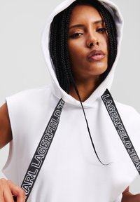 Balta bevandenė su dideliu kapišonu, puošta juodomis prekės ženklo juostelėmis. Lygus tekstūras su minimaliais dizaino akcentais.