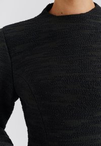 Schwarzes Texturkleid mit gewebtem Muster, Rundhalsausschnitt und figurbetontem Design, das unterschiedliche Stoffstärken zur Geltung bringt.