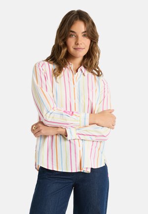 Woman with wavy brown hair wearing a pastel vertical striped button-up shirt and dark blue jeans, standing with arms crossed.