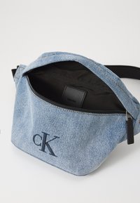 Borsa in vita in denim dalla forma curva, colore azzurro chiaro, con interno nero, chiusura a zip e logo "CK" ricamato sul davanti.