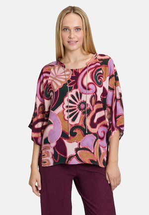 Mehrfarbige Blumenshirt mit weitem Ausschnitt, Dreiviertelärmeln und lockerer Passform. Es zeigt Muster in Rosa-, Lila- und Brauntönen.