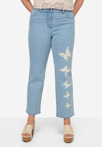 Vaqueros de mezclilla azul claro con corte recto y bordado de mariposa a lo largo de la pierna izquierda. Combinados con sandalias de plataforma beige.