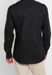 Svart långärmad skjorta i mjukt tyg med knäppningar vid ärmsluten och böjd nederkant. Har en klassisk krage och minimalistisk design.