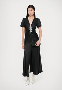 Juoda satino suknelė su trumpomis, platėjančiomis rankovėmis ir gilia V formos iškirpte, juosmenyje puošta papuošalu. Modelis laiko juodą rankinę.