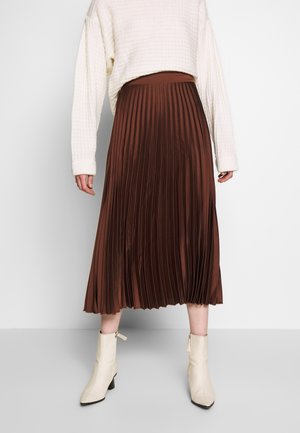 Osoba ubrana w biały sweter o fakturze, brązową plisowaną spódnicę midi oraz kremowe botki, stojąca na jednolitym tle.