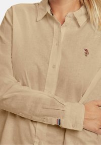 Camicia beige a maniche lunghe con design a bottoni, caratterizzata da un piccolo logo ricamato sul petto e un'accattivante striscia sul polsino.