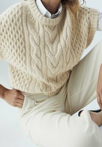 Stickad beige tröja med strukturmönster i flätor, korta ärmar och rund halsringning, ihop med ljusgräddfärgade ribbade byxor.