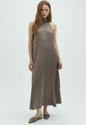 Femme portant une robe longue taupe sans manches avec un col montant et des ballerines slingback à bout pointu, debout devant un fond clair uni.