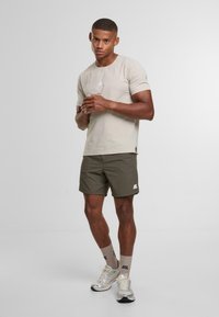 Uomo muscoloso in piedi con una maglietta beige, pantaloni corti oliva, calzini beige e scarpe da corsa argentate su uno sfondo chiaro e semplice.