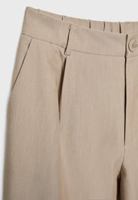 Pantalon beige en tissu lisse et texturé, avec des plis à l'avant, une ceinture et une fermeture par bouton.