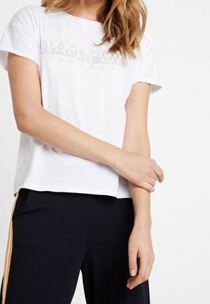 Nainen, jolla on päällään valkoinen lyhythihainen Napapijri-t-paita ja mustat housut, joissa on keltaiset raidat, pitää vasemmasta ranteestaan oikealla kädellään.