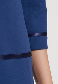 Blått tyg med en slät textur, prydd med mörkblå satängband vid ärmarna, vilket framhäver en strukturerad design.