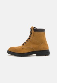 Bottines en suede marron avec un col en cuir foncé, bout arrondi, fermeture à lacets à l'avant et semelle en caoutchouc robuste. Détails de couture contrastants présents.