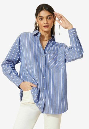 Camicetta a righe blu con righe verticali bianche, dotata di taschino sul petto e vestibilità comoda. Indossata con pantaloni chiari.