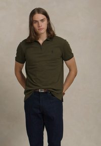 Polo verde escuro com mangas curtas e colarinho com fecho éclair, combinado com jeans azul escuro. Design simples, material em algodão e sem padrões.