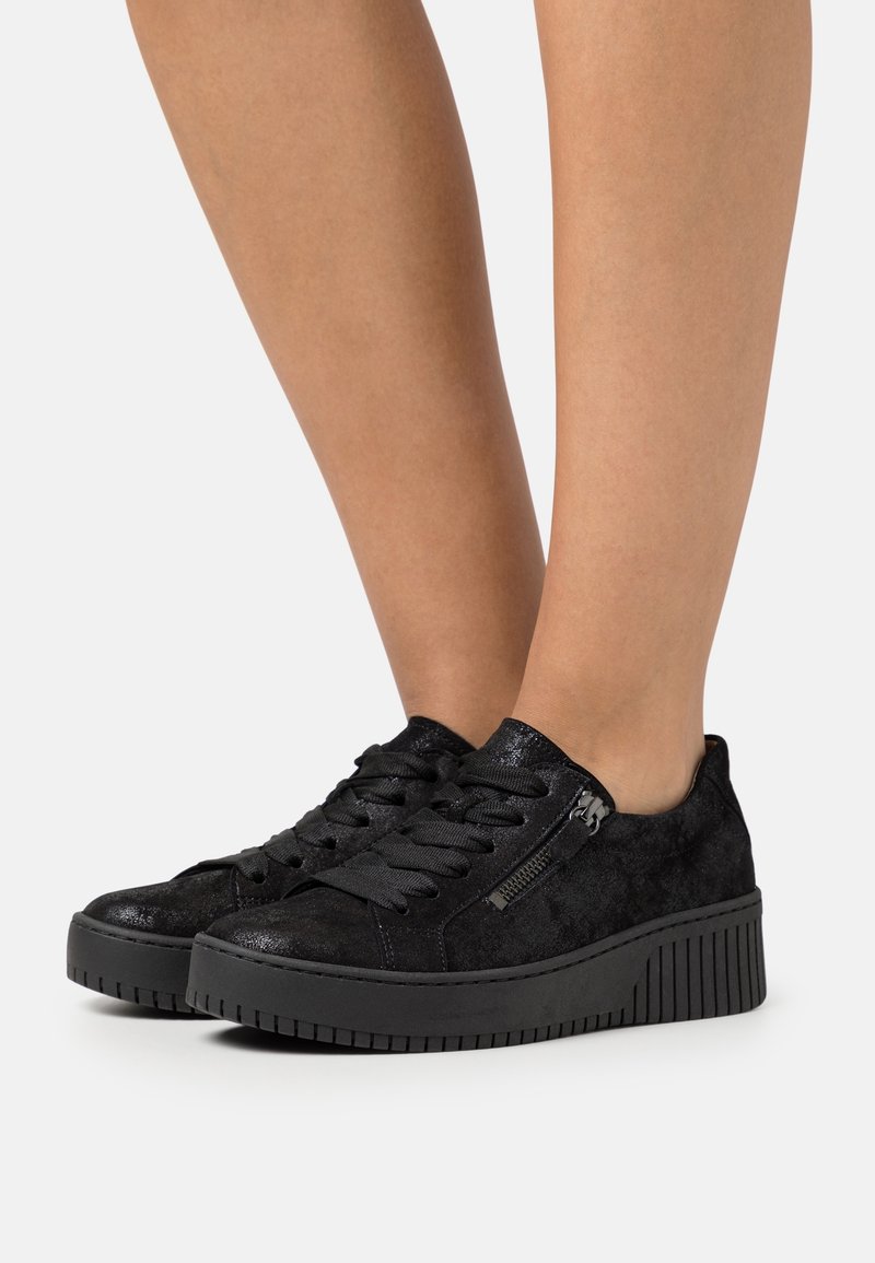 Personne portant des baskets en daim noir avec semelles épaisses, larges lacets et fermetures éclair latérales, debout sur un fond clair et uni.