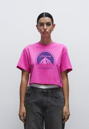 T-shirt corta rosa brillante con un logo circolare blu scuro di Paramount e una grafica di montagna, abbinata a jeans grigi a vita alta.