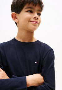Ragazzo con capelli corti e scuri che indossa una maglia a maniche lunghe blu scuro, sorridendo leggermente e guardando di lato con le braccia incrociate.