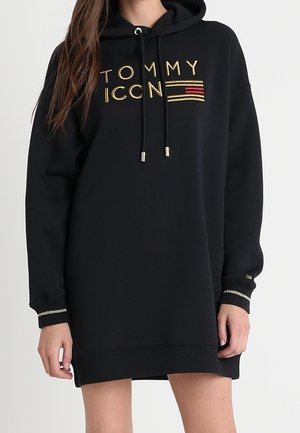 Czarna oversize sukienka z kapturem z złotym napisem "TOMMY ICON" i haftem flagi, srebrne mankiety na rękawach, noszona przez stojącą osobę.