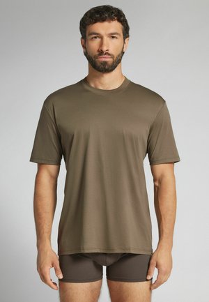 Mężczyzna z brodą, ubrany w jednokolorowy brązowy t-shirt z krótkim rękawem i ciemnobrązowe dopasowane szorty, stojący na szarym tle.