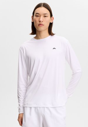 Joven con cabello liso hasta los hombros lleva una camisa blanca de manga larga y pantalones blancos sobre un fondo liso.