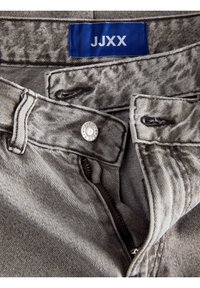 Grå denimjeans med en silverfärgad metalldragknapp och dragkedja, försedd med en blå etikett med texten "JJXX" sydd på midjebandet.