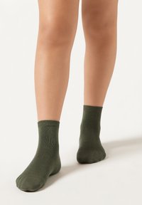 Grønne ankelstrømper lavet af struktureret stof med ribbede toppe og en glat finish. Vist på ben mod en neutral baggrund.