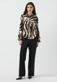 Langermet bluse med et svart og kremfarget abstrakt stripe mønster, med en knappekrage og vide ermer, kombinert med svarte bukser.
