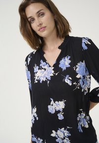 Blusa nera con motivo floreale blu e bianco, caratterizzata da scollatura a V e colletto arricciato, maniche lunghe con polsini arricciati e tessuto leggero.