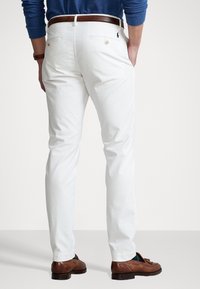 Białe bawełniane chinosy o dopasowanym kroju, z tylnymi kieszeniami i brązowym skórzanym paskiem. Noszone z brązowymi butami.