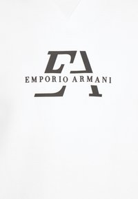 Bílý tričko s V-výstřihem a hladkou texturou, s černým logem "EA" a textem "EMPORIO ARMANI" umístěným na přední straně uprostřed.