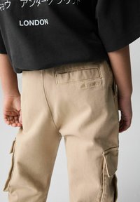Beige cargobyxor gjorda av kraftigt tyg, med sidfickor, en bakficka och sydd branding i en subtil, präglad design.