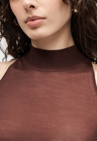 Γυναίκα με καστανά σγουρά μαλλιά που φορά χρυσό κρίκο σκουλαρίκι και αμάνικη καφέ μπλούζα με μεγάλο λαιμό, που κόβεται στο πηγούνι και στους ώμους.