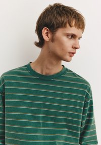 Tricou cu mâneci lungi cu dungi verzi, confecționat din țesătură moale. Are dungi subțiri albe și portocalii, guler rotund cu nervuri și croială lejeră.